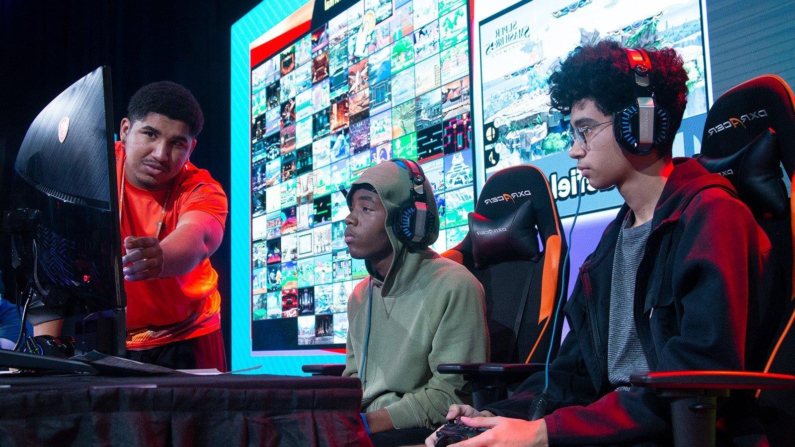 Two Florida high school students seated on stage in front of a gaming monitor with a Full Sail Armada athlete helping them to their right.