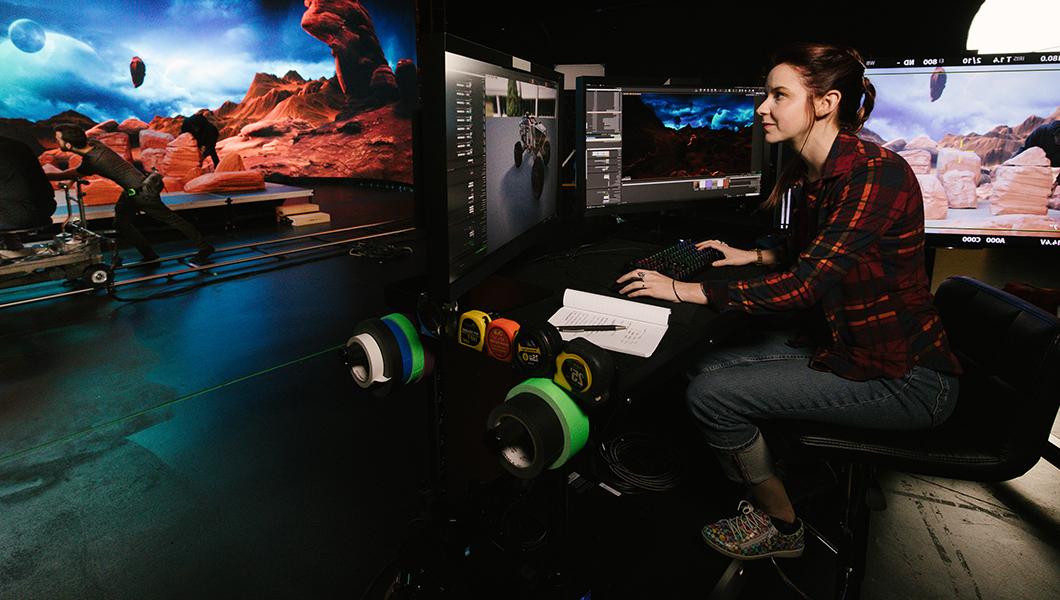 A woman is sitting at a desk in front of several monitors. She is in a virtual production studio that has been designed to look similar to Mars.