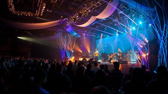 A band plays music in a large, 黑暗的礼堂里有几盏明亮的蓝色和紫色的灯指向舞台.