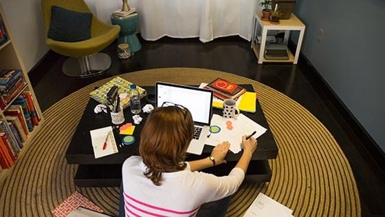 一个学生盘腿坐在咖啡桌前的地板上，手里拿着一台笔记本电脑, 论文, 笔记本电脑, 和笔.