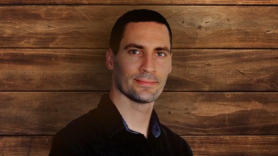 Justin Hosie standing in front of a paneled wood background. He is wearing a dark denim shirt and gently smiling.