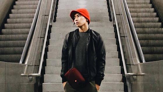 A young Asian man wearing a red knit cap, black jacket and shirt, green pants, and a burgundy bag stands between two escalators. His hands are in his pockets and he is looking off to the left.