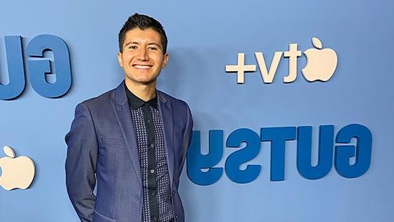 A person with short, dark hair is smiling while wearing a blue suit against a light blue step and repeat that shows the logos for ‘Gutsy’ and Apple TV+.