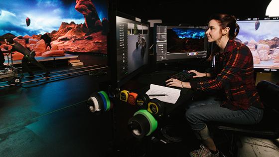 A woman is sitting at a desk in front of several monitors. 她在一个虚拟的制作工作室里，这个工作室被设计得看起来和火星很像.