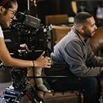 Behind the scenes on a student film set, 一个学生坐在椅子上拿着笔记本，另一个学生在胶片相机后面工作