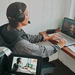 Grad Sydney Phillips, 一个深棕色头发的女人，穿着灰色连帽运动衫，戴着黑色耳罩式耳机, 坐在写字台前的笔记本电脑前的记者包厢里俯瞰着足球场上穿着制服的运动员.