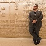 Wearing a suit 和 glasses, Stephen Beres leans against a beige, textured stone wall with the HBO logo carved into it.