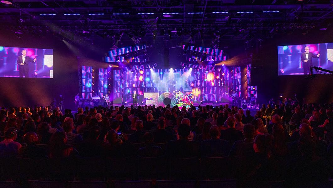 一大群人坐在增高的舞台前. 在紫色的屏幕前，一个巨大的彩色字母“HOF”雕塑坐落在舞台顶端. 一名身穿黑色西装的男子站在舞台上，并在舞台旁边的屏幕上拍照.