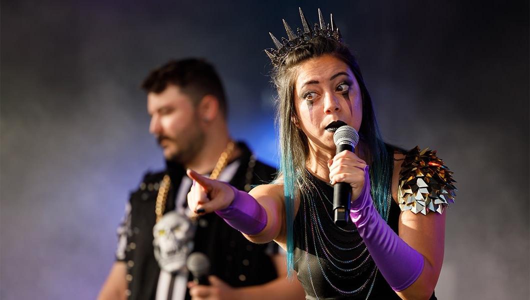 A woman in a metal crown and stage makeup pointing outward while making an exaggerated face at the crowd.