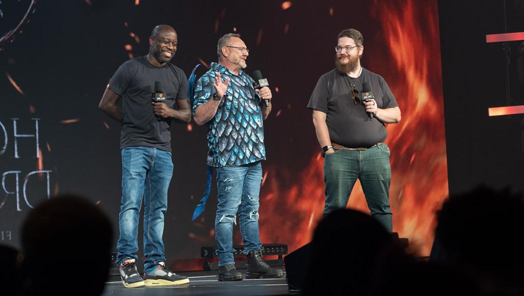 On stage inside the Full Sail Live Venue, Stephen Beres, Rick Ramsey, and Leslie Brathwaite hold mics and stand before a large screen showing the fiery House of the Dragon logo.